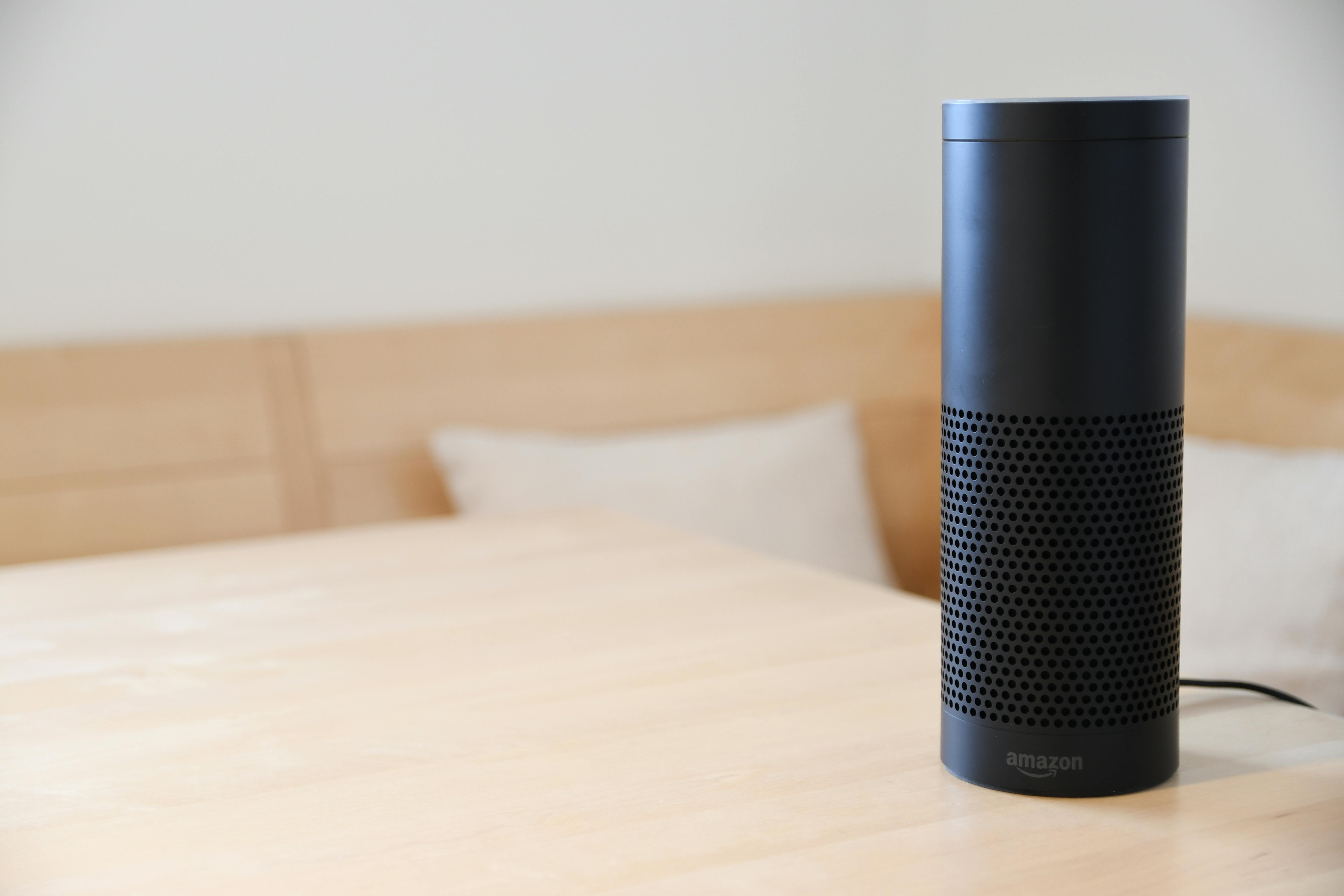 A black smart speaker resting on a light-colored wooden table in a cozy indoor setting.